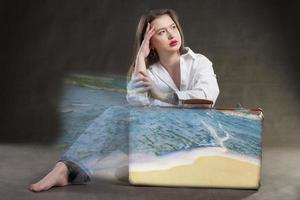 A beautiful girl sits with an old suitcase from which the ocean wave flows. photo