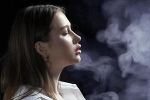 Profile of a young female model with perfect sensual lips on a black background in puffs of smoke. Beautiful facial skin, augmentation of female lips, glamorous elegance. photo