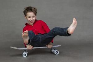 gracioso pequeño chico sentado en un Clásico patineta descalzo en un gris antecedentes. foto