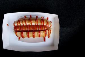 Hot dog in the slit of a partially sliced bun and top with tomato sauce, mayonnaise put in white paper box for take home and black background. photo