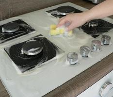 Cleaning dirty gas stove, foam from household chemicals. Hands hold a washing sponge in the foam. White glass hob surface. The concept of household chores, house cleaning. photo