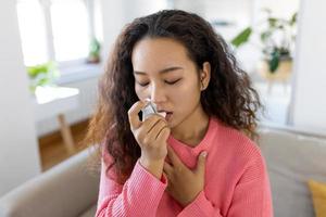 asiático mujer utilizando inhalador mientras sufrimiento desde asma a hogar. joven mujer utilizando asma inhalador. de cerca de un joven asiático mujer utilizando asma inhalador a hogar. foto