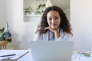 youn asiático médico utilizando ordenador portátil y sentado a escritorio. mujer profesional médico médico vistiendo blanco Saco y estetoscopio trabajando en computadora a lugar de trabajo. foto