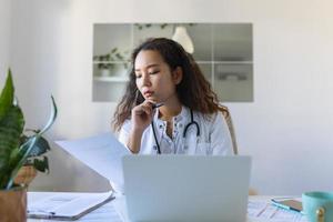 youn asiático médico utilizando ordenador portátil y sentado a escritorio. mujer profesional médico médico vistiendo blanco Saco y estetoscopio trabajando en computadora a lugar de trabajo. foto