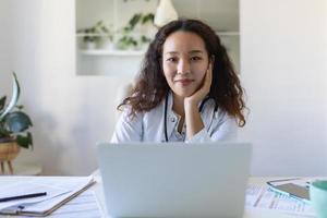 youn asiático médico utilizando ordenador portátil y sentado a escritorio. mujer profesional médico médico vistiendo blanco Saco y estetoscopio trabajando en computadora a lugar de trabajo. foto