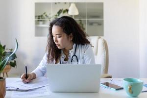 youn asiático médico utilizando ordenador portátil y sentado a escritorio. mujer profesional médico médico vistiendo blanco Saco y estetoscopio trabajando en computadora a lugar de trabajo. foto