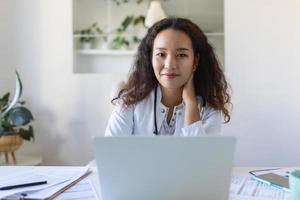 youn asiático médico utilizando ordenador portátil y sentado a escritorio. mujer profesional médico médico vistiendo blanco Saco y estetoscopio trabajando en computadora a lugar de trabajo. foto