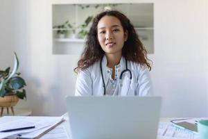 youn asiático médico utilizando ordenador portátil y sentado a escritorio. mujer profesional médico médico vistiendo blanco Saco y estetoscopio trabajando en computadora a lugar de trabajo. foto