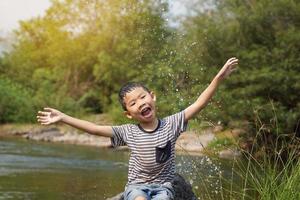 Asian boy taking a break from playing in a stream Sit on the rock, smiling brightly, arms outstretched to enjoy the splashing water happily. soft and selective focus. photo