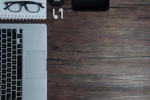 wooden office desk with laptop, blank notebook, cordless earphones and glasses, dark background, wooden background, Top view with copy space, empty space. photo