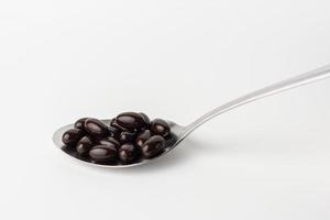 Black soft gel capsules in a stainless steel spoon on a white background. Vitamin food supplements photo