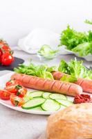 Ready to eat fried sausages, chopped tomatoes and cucumber vegetables on a plate and a bun on the table. Hearty homemade snack. Vertical view photo