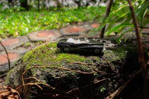 A photo after some edits. A black toy car on a mossy stone. A concept of go to nature with classic car.