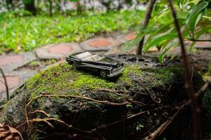 A photo after some edits. A black toy car on a mossy stone. A concept of go to nature with classic car.