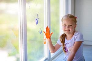 niños niña dibujar con palmas en el ventana. pintado manos salir un marca en el vaso. cuarentena. permanecer hogar. foto