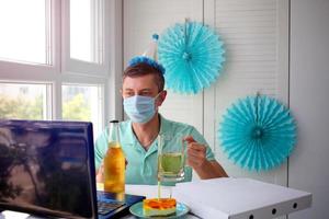 A man in a medical mask sits in front of a laptop, celebrates a birthday with friends over the Internet and holds a glass of alcohol in his hands. photo