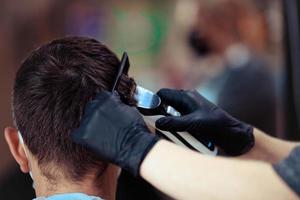 un peluquero con seguridad medidas para COVID-19, sostiene un maquinilla de afeitar y un Corte de pelo para un hombre, cortes su pelo con caucho guantes en un belleza salón foto