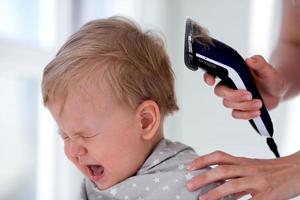 Female hands trim a crying baby with an electric hair clipper in a hairdresser. First haircut. photo