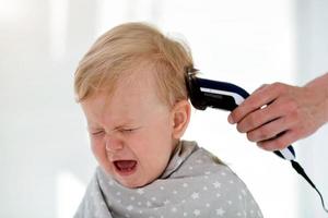 Female hands trim a crying baby with an electric hair clipper in a hairdresser. First haircut. photo