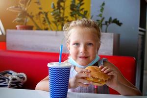 un pequeño niña se sienta en un café en un médico máscara y sostiene un hamburguesa y soda en su manos. apertura un cafetería. trabajo café con seguridad medidas para COVID-19. foto