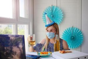 un mujer en un médico máscara se sienta en frente de un computadora portátil, celebra cumpleaños con amigos terminado el Internet, y bebidas alcohol. cumpleaños en línea foto
