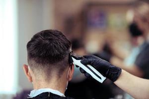 un peluquero con seguridad medidas para COVID-19, sostiene un maquinilla de afeitar y un Corte de pelo para un hombre, negociaciones a un distancia, cortes su pelo con caucho guantes foto