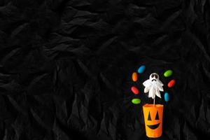 Handmade paper cup in the form of a pumpkin, a confession and jelly candies on a black crumpled background. Top view . photo