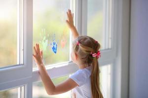niños niña dibujar con palmas en el ventana. pintado manos salir un marca en el vaso. cuarentena. permanecer hogar. foto