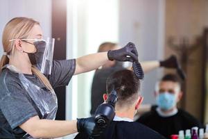 peluquero con seguridad medidas para COVID-19, golpe a secas un hombre en un medicina mascarilla, social distancia, corte pelo con un médico mascarilla, ojo máscara y caucho guantes en un belleza salón foto