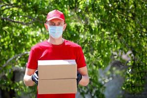 Courier in a red T-shirt and a cap in a medical mask and gloves holds a cardboard box in your hands, outdoor. photo