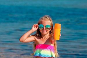 Beautiful little girl in a swimsuit and sunglasses stands by the sea and holds a sunscreen in her hands. photo