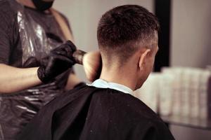 un peluquero con seguridad medidas para COVID-19, sostiene tijeras en su manos y cortes un hombre, corte pelo con caucho guantes foto