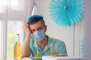 A sad man celebrates a birthday alone in a medical mask and looks at the camera. . A man in a festive cap sits at a table with beer, pizza and cake. photo