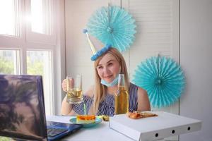 un mujer en un médico máscara se sienta en frente de un computadora portátil, celebra cumpleaños con amigos terminado el Internet, y bebidas alcohol. foto