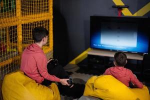 Two brothers playing video game console, sitting on yellow pouf in kids play center. photo