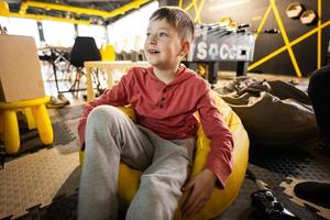 Boy watching tv while playing video game console, sitting on yellow pouf in kids play center. photo