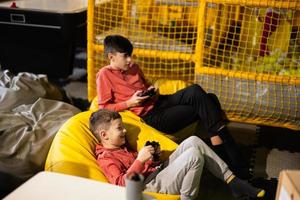 Two brothers playing video game console, sitting on yellow pouf in kids play center. photo