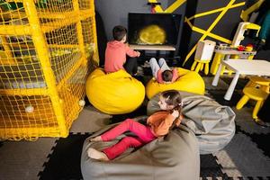 Tres niños jugando vídeo juego consola, sentado en amarillo puff en niños jugar centro. foto