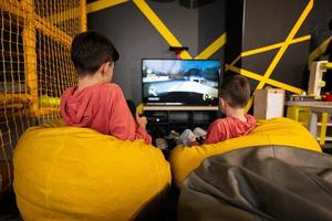 Two brothers playing race video game console, sitting on yellow pouf in kids play center.