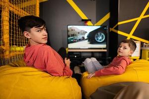 Two brothers playing race video game console, sitting on yellow pouf in kids play center.