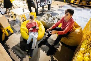 Two brothers playing video game console, sitting on yellow pouf in kids play center. photo