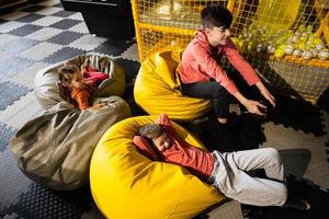 Tres niños jugando vídeo juego consola, sentado en amarillo puff en niños jugar centro. foto