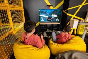 Two brothers playing race video game console, sitting on yellow pouf in kids play center. photo