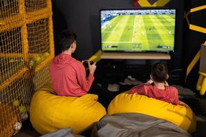 Two brothers playing football video game console, sitting on yellow pouf in kids play center. photo