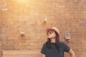 joven bonito alegre niña posando con el pared a soleado día, teniendo divertido solo, elegante Clásico ropa sombrero y Gafas de sol, foto