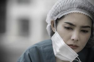 Tired depressed female asian scrub nurse wears face mask blue uniform sits on hospital floor,Young woman doctor stressed from hard work photo