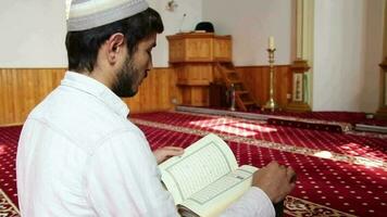 un joven hombre lee el Corán en pie en el mesa por siguiendo eso con su dedo, un adolescente con un religioso sombrero en su cabeza leyendo el Corán en el mezquita video