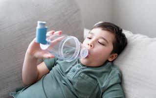 enfermo niño utilizando asma inhalador para alergias, pobre chico cansado desde cofre tos participación inhalador espaciador, probado niño teniendo asma alergia foto