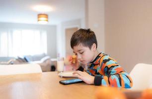 sano niño teniendo desayuno, contento niño chico utilizando móvil teléfono mirando, leyendo o jugando juegos un mientras comiendo alimento, colegio niño cuerno y Leche antes de Vamos a colegio en el Mañana foto