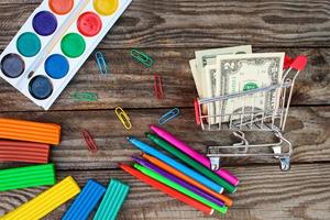 Shopping cart with money. Office and school supplies on old wooden background. Concept is to buy stationery objects. photo